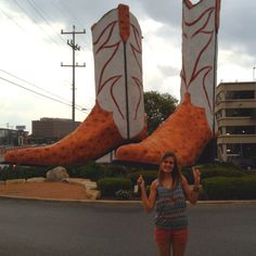 a woman standing in front of two giant boots on the side of a road with her hands up