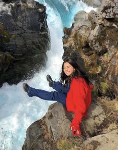 a woman sitting on the edge of a cliff next to a river with water rushing down it