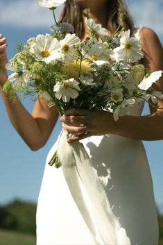 a woman in a white dress holding a bouquet of daisies and wildflowers