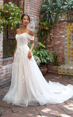 a woman standing in front of a brick wall wearing a wedding dress with an off the shoulder design