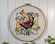 a cross - stitch butterfly with yellow flowers in a frame on a white wall next to a potted plant