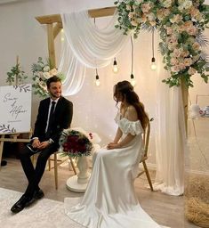 a man and woman sitting on chairs in front of a white backdrop with floral arrangements