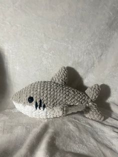 a crocheted stuffed shark laying on top of a white bed sheet with black eyes