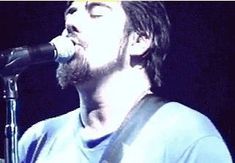 a man singing into a microphone while holding a guitar in front of his face and wearing a blue shirt