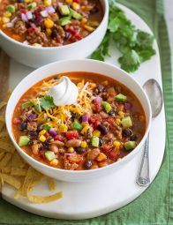 two white bowls filled with chili and corn on top of a green place mat next to silverware