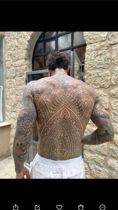 the back of a man with tattoos on his chest and arms, standing in front of a stone building