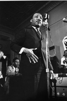 a black and white photo of a man speaking into a microphone in front of a group of people