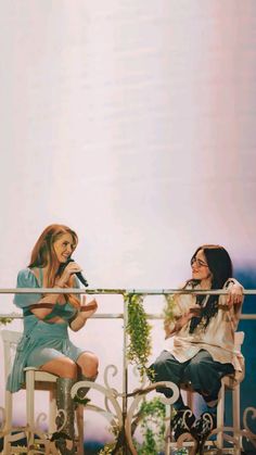 two women sitting on chairs talking to each other in front of a balcony overlooking the ocean