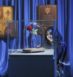 a woman standing in front of a glass case with a flower inside it and blue curtains behind her