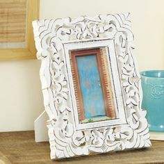 an ornate white frame sitting on top of a wooden table next to a blue mug