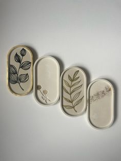 three small trays with designs on them are lined up against a white wall, one is empty and the other has leaves painted on it