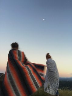 two women wrapped in blankets are walking on a hill at sunset with the moon behind them