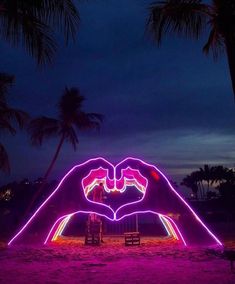 two hands making a heart shape in front of palm trees at night with purple light