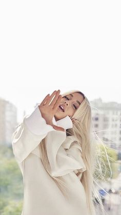 a woman with long blonde hair is holding her hand up to her face while standing in front of a window