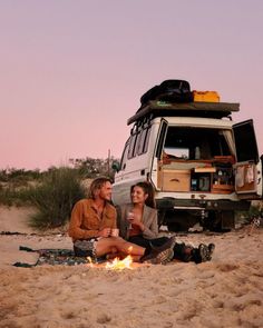 two people sitting in the sand next to a camper van with a tent on top