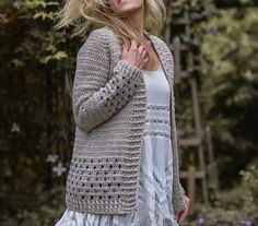 a woman is walking in the woods wearing a crochet cardigan and white dress