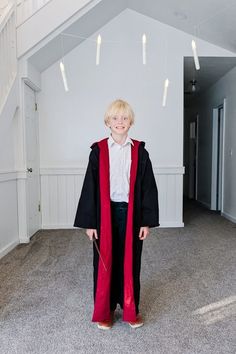 a young boy wearing a graduation gown and holding a wand in an empty room with stairs leading up to the second floor