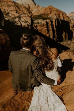 a bride and groom are sitting on the edge of a cliff looking at the mountains