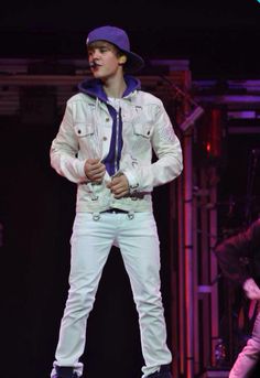 a man standing on top of a stage wearing white pants and a blue hat with his hands in his pockets