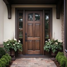 two planters with white flowers are in front of a brown door and brick walkway