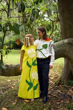 two people standing next to each other in front of a tree with leaves on it