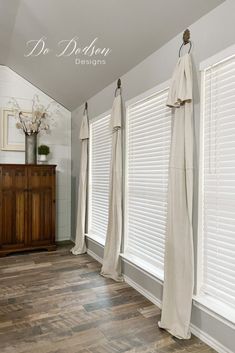 a room with white curtains and wood flooring in front of two windows that have drapes hanging from them