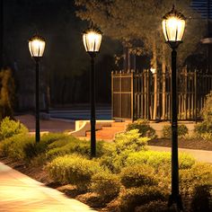 three street lamps are lit up in the evening on a sidewalk next to bushes and trees