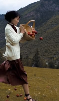 a woman holding a basket full of apples in her hand while walking through an open field