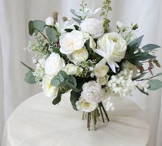 a vase filled with white flowers and greenery on top of a round table in front of a white curtain