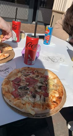 a pizza sitting on top of a white table next to two sodas and cups