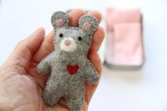 a hand holding a small gray teddy bear with a heart on it's chest