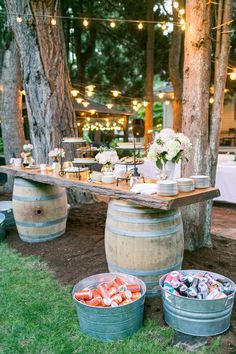 an outdoor table with wine barrels and food on it, surrounded by string lights in the background