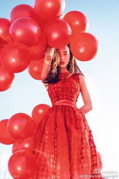 a woman in a red dress is surrounded by many red balloons that are floating above her head