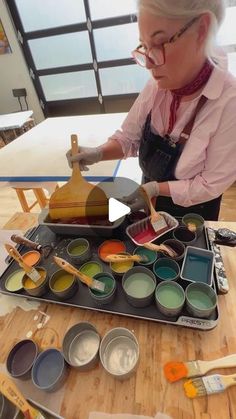 an older woman is painting on a table with paint cans and brushes in front of her