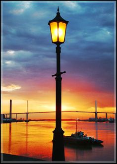 a street light sitting on the side of a body of water at sunset with a bridge in the background