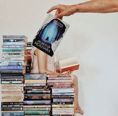 a stack of books sitting on top of a person's lap next to a white wall