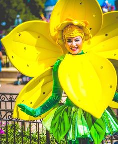 a woman dressed as a yellow flower with green leaves on her head and arms, standing in front of a fence