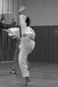 two people doing tricks in an indoor gym area, one holding a laptop and the other standing up