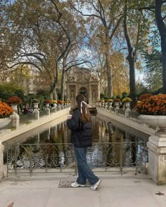 a woman standing in front of a pond surrounded by trees and orange flowers with a fountain behind her