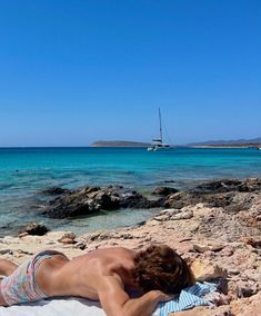 a man laying on top of a sandy beach next to the ocean with a sailboat in the background