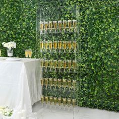 a table topped with lots of champagne glasses next to a wall covered in greenery