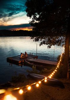 two people sitting on a dock at night with lights strung across the water and boats in the background