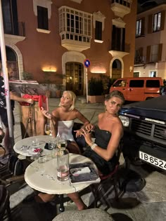 two women sitting at an outdoor table with drinks in front of some cars and people