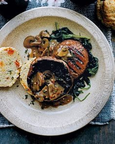 a white plate topped with meat covered in mushrooms and spinach next to an english muffin