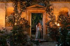 a woman standing in an open door to a building with candles on the outside and greenery around her