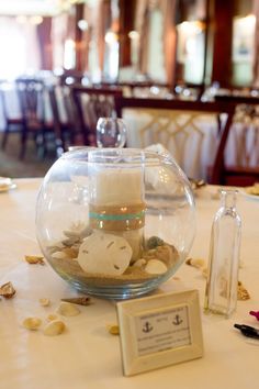 a fish bowl filled with sand and sea shells on top of a white table cloth