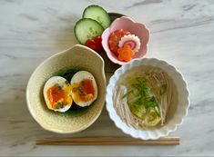 two bowls filled with food next to chopsticks and cucumbers on a table