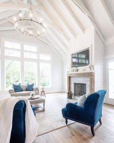 An elegant farmhouse living room with a vaulted white shiplap ceiling and shiplap walls with a built-in TV above a wood beam mantel and brick fireplace. A wagon wheel chandelier hangs in the center of the room and large windows bathe the room with natural light...   Image: the.old.barn