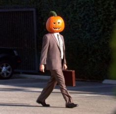 a man in a suit walks down the street with a pumpkin on his head