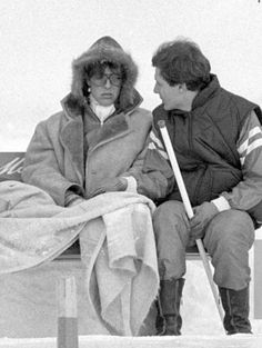 two people sitting on a bench in the snow, one holding a stick and the other looking at each other